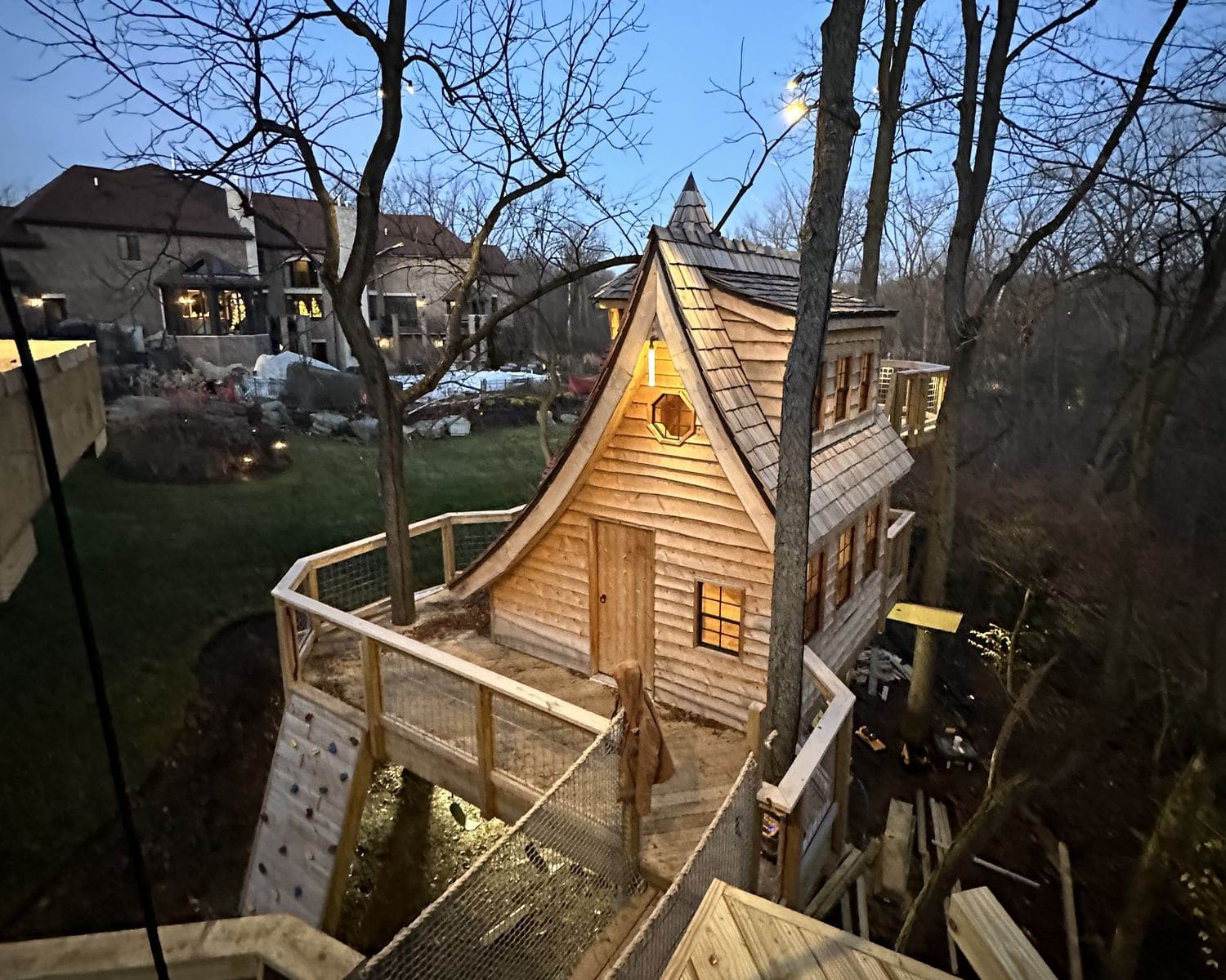 A custom residential treehouse at dusk, curved cedar-shake gable and lit windows